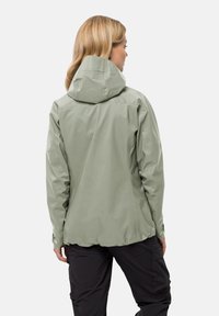 Giacca impermeabile verde chiaro con cappuccio, caratterizzata da una texture liscia e orlo regolabile. Il design è aderente e privo di hardware visibile.