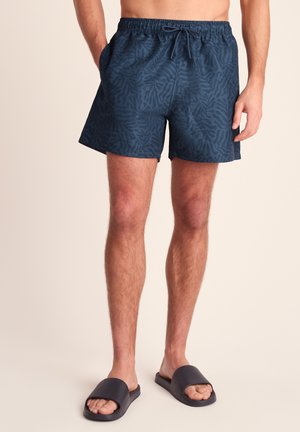 Man wearing dark blue patterned swim shorts and black slide sandals stands with hands in pockets against a plain background.