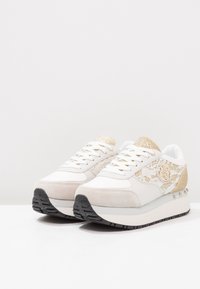 Hvide sneakers med snørebånd og design med guldaccenter, ruskinds-overlays, flade snørebånd og en struktureret sort sål for greb og stabilitet.