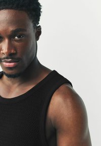 Zwarte gebreide tanktop met een gestructureerd patroon; heeft brede bandjes en een ronde halslijn. Het model heeft kort, krullend haar en staat tegen een witte achtergrond.