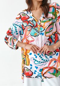 Blouse multicolore avec col en V, manches trois-quarts, et motifs floraux abstraits en rouge, bleu, rose et vert sur fond blanc.