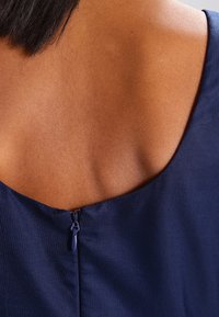 Marineblauwe mouwloze jurk met een ronde hals aan de achterkant, voorzien van een ritssluiting. Glad textuur met subtiele geribbelde stofdetails.