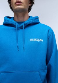 Blauwe katoenen hoodie met een kangoeroezak, capuchon met trekkoord en een wit logo op de linkerborst. Gladde textuur en relaxte pasvorm.