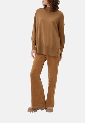 Brauner Strickpullover mit hohem Kragen und seitlichen Schlitzen, kombiniert mit braunen, ausgestellten Cordhosen und hellbraunen Plateau-Schuhen.