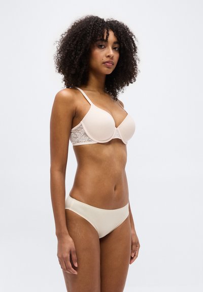 Γυναίκα με σγουρά μαλλιά, που φοράει μπεζ σουτιέν με δαντελωτές πλευρές και ασορτί άνετο εσώρουχο, στέκεται μπροστά σε απλό φωτεινό φόντο.