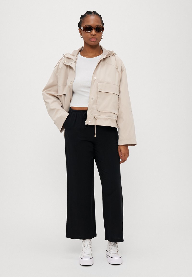 Veste courte beige clair avec poches avant, ourlet à cordon de serrage, associée à un pantalon noir à jambes larges et des baskets montantes blanches. Lunettes de soleil marron incluses.