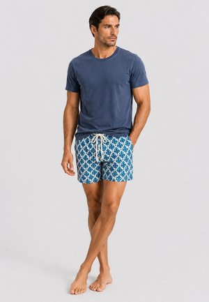 Mann steht barfuß, trägt ein marineblaues T-Shirt und blaue, gemusterte Kordelzugshorts, blickt nach rechts und hat die Hände in den Taschen.