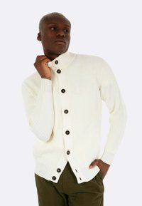 Cardigan bianco a coste con colletto alto e bottoni scuri. Presenta spacchi laterali e tessuto testurizzato. Indossato sopra pantaloni verde oliva.