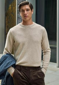 Beige gebreide trui met een ronde hals, gecombineerd met donker corduroy broek. Het model houdt een blauwe jas over één arm gedrapeerd.
