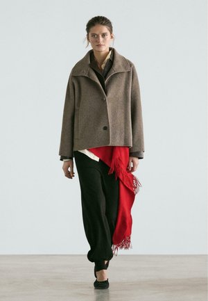 Cappotto in lana marrone con collo e chiusura a bottoni, sovrapposto a un completo nero, accentuato da una sciarpa rossa con frange. Tessuto liscio, vestibilità rilassata.
