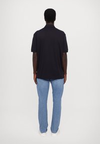 Svart polo skjorte med korte ermer, avslappet passform, iført med lyseblå jeans og hvite joggesko, vist bakfra mot en enkel bakgrunn.