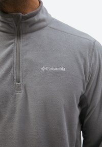 Man wearing light grey Columbia fleece jacket with quarter zip and logo on the chest against plain background.