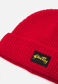 Căciulă tricotată roșie cu un model texturat și un etichetă neagră cu logo-ul auriu "Stan Ray". Designul este simplu și funcțional.