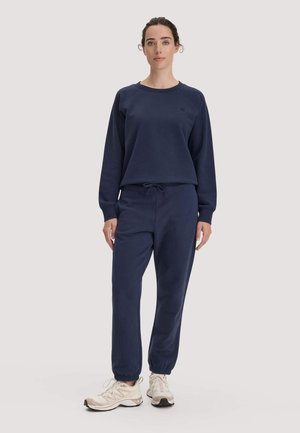 Frau steht und trägt einen marineblauen Sweatshirt und Jogginghose mit beigen Sportschuhen auf einem schlichten hellen Hintergrund.