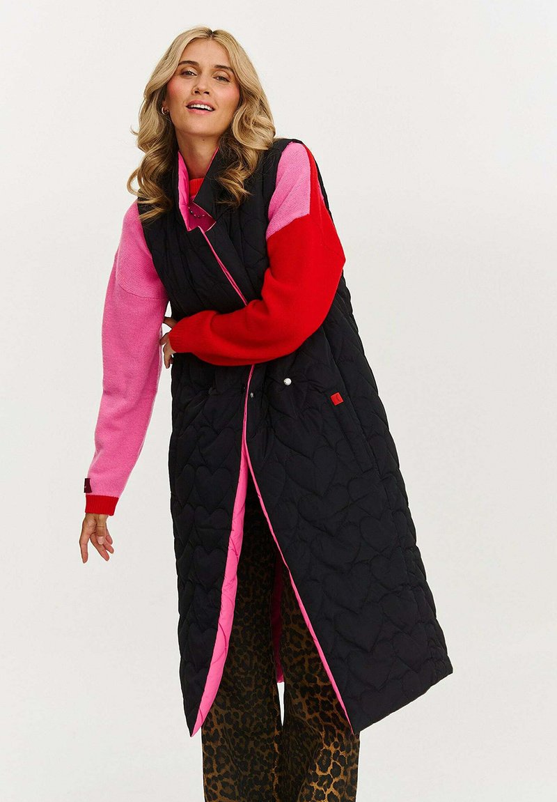 Czarny pikowany długi kamizelka z różowym podbiciem, kapturem i serduszkowym wzorem. Noszona na różowym i czerwonym swetrze oraz spodniach w panterkę.