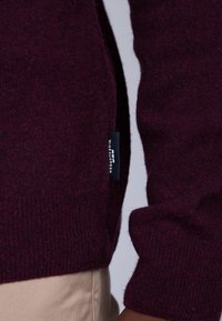 Le pull en tricot bordeaux présente une texture côtelée, des manches ajustées et une étiquette latérale affichant "Miseaugreen". Matériau doux et chaud.