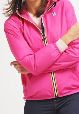 Vrouw draagt een felroze jas met ritssluiting, elastische manchetten en gestreepte ritssluitingdetails, staand met gekruiste armen.