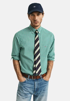 Camisa de cuadros verde y blanca, corbata navy a rayas, vaqueros azules, cinturón marrón y gorra navy. El modelo tiene el cabello corto y tatuajes.