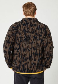 Giacca in pile marrone con motivo astratto nero, design con colletto, superficie testurizzata e vestibilità rilassata. Indossata sopra un indumento giallo.