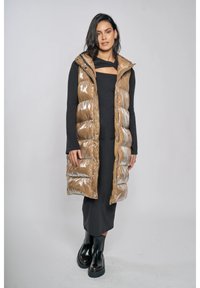 Lange, glanzende bruine donsbodywarmer met capuchon, met doorgestikte secties en zijzakken, gedragen over een zwarte jurk met lange mouwen en stevige laarzen.