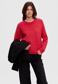 Rood gebreid sweater met een ronde hals en lange mouwen, gecombineerd met een donkere jas. De sweater heeft een zachte, gestructureerde uitstraling.