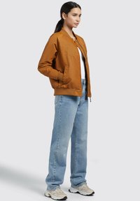 Giacca bomber marrone con dettagli ricamati, chiusura con zip e tasche laterali, abbinata a jeans a gamba dritta blu chiaro e sneakers beige.