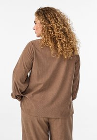 Brown, ribbed long-sleeve top with a relaxed fit and rounded hem, featuring loose cuffs. The textured fabric adds depth to the design.