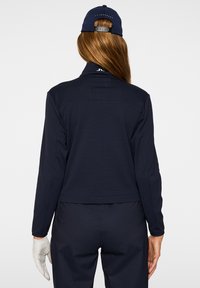 Camicia a maniche lunghe blu navy con colletto, tessuto leggero, design leggermente crop e dettaglio di pieghe sul retro. Abbinata a un cappellino blu navy e guanti bianchi.