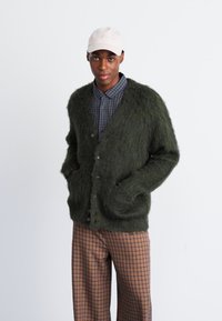 Πράσινο φανελάκι με υφή, υπερμεγέθες σχέδιο και μπροστινές τσέπες. Φοριέται πάνω από πουκάμισο καρό και συνδυάζεται με καρό παντελόνι.