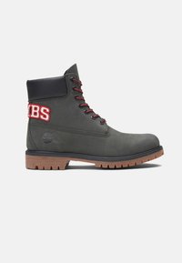 Botín de ante gris con detalles en negro, cordones rojos y parche bordado "JBS". Suela robusta para tracción, presenta un diseño de punta redonda.
