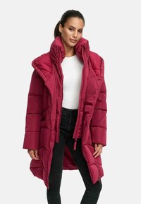 Ilgs, pūkains mētelis spilgtā bordo krāsā. Iespējamais augsts apkakle, rāvējslēdzēja aizdare un quilted dizains ar horizontālu šuvēm. Kombinēts ar melniem biksēm.