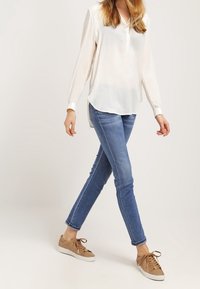 Osoba kráčející v bílé blůze s dlouhým rukávem, modrých skinny džínách a hnědých šněrovacích teniskách na jednoduchém bílém pozadí.