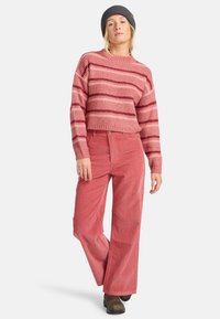Gestreifter pinker Strickpullover mit breitem Rippenmuster, kombiniert mit hochgeschnittenen korallenfarbenen Cordhosen. Die Accessoires umfassen eine dunkelgraue Mütze.