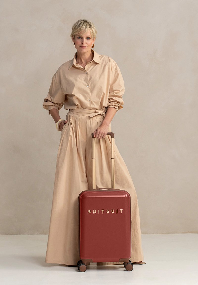 Een jonge vrouw met kort blond haar staat met een rode koffer met "SUITSUIT" erop geschreven, gekleed in een beige oversized shirt en rok.