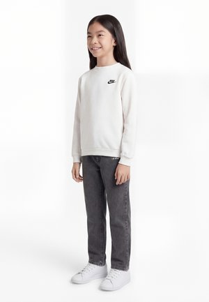 Niña sonriente con cabello largo y negro, vistiendo una sudadera blanca de Nike, jeans gris oscuro y zapatillas blancas, de pie con una postura relajada.