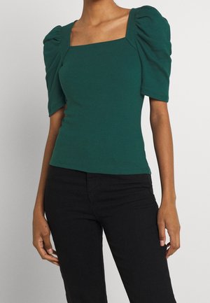 Donna che indossa un top aderente verde scuro con maniche a sbuffo corte e scollatura quadrata, abbinato a pantaloni neri.