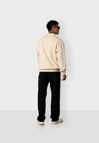 Cremiges, übergroßes Sweatshirt mit gerippten Bündchen, kombiniert mit schwarzen Hosen und weißen Sneakers, von hinten betrachtet, vor einem hellen Hintergrund.