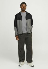 Giacca a zip nera e grigia con cappuccio, abbinata a una maglietta grigia e pantaloni scuri a vestibilità rilassata, con polsini elasticizzati e tasche laterali.