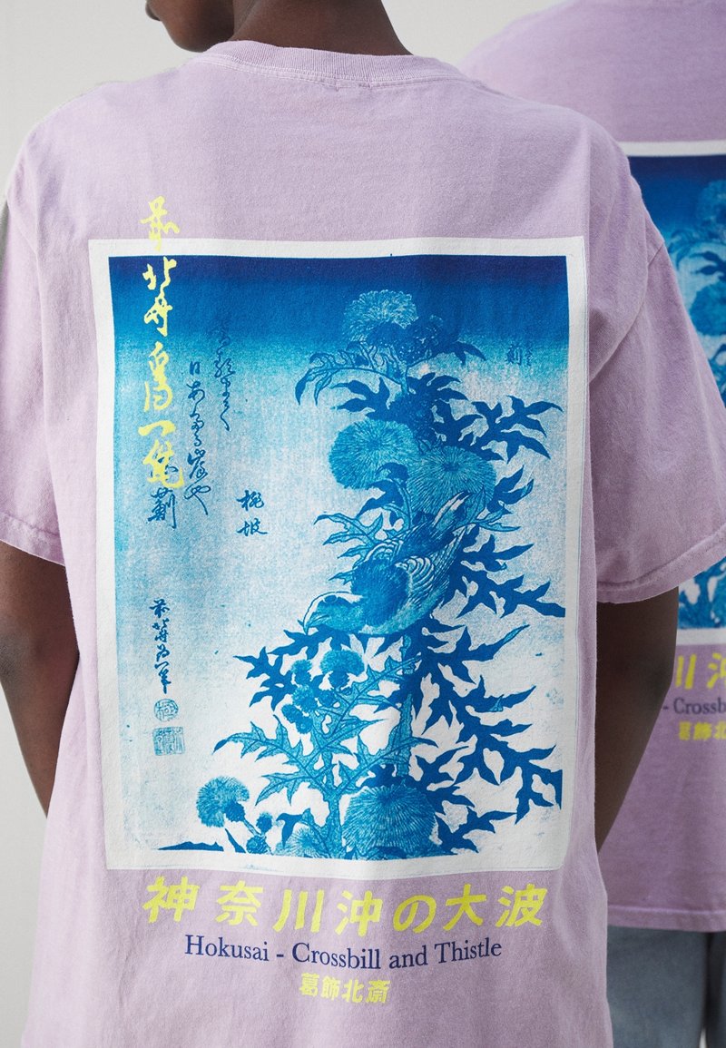 Lavendel bomull t-shirt med ett grafiskt tryck av törne och en fågel i blått och vitt, med gult text som gör designen.
