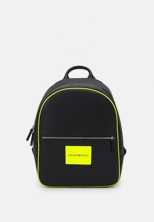 Musta Emporio Armani reppu, jossa on neonkeltaiset yksityiskohdat, etutasku vetoketjulla ja neonkeltaisen värinen logopatchi valkoista taustaa vasten.