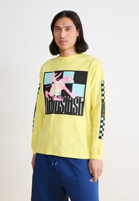 T-shirt jaune à manches longues présentant un grand graphique noir et rose d'un cow-boy sur un cheval. Détails en chevrons sur les manches. Matériau en coton doux.