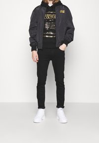 Osoba v černé bomberce přes černou mikinu s lesklým zlatým potiskem, černých slim kalhotách a bílých chunky teniskách na jednobarevném pozadí.