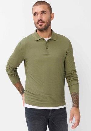 Man met kort haar en baard, draagt een olijfgroene polo met lange mouwen en zwarte spijkerbroek, kijkt licht omhoog naar links.