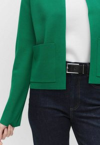 Grön kortärmad cardigan med en texturerad yta och två stora framfickor, i kombination med mörk denimjeans och en vit topp.