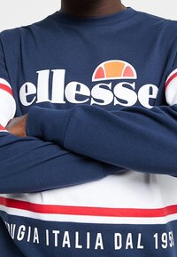 Person som bär en blå, vit och röd ellesse-tröja med korsade armar och texten "Perugia Italia Dal 1959" på nederdelen.