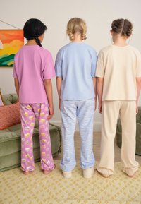 Tre bambini sono visti di spalle, indossano magliette pastello: rosa, azzurre e beige. I pantaloni del pigiama presentano motivi di torte e gatti. Si possono notare texture morbide.