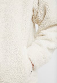Cremefarvet fleece-pullover med en tekstureret overflade, dropped shoulder-seams og sidelommer for ekstra funktionalitet.