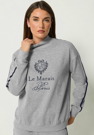 Kvinde med langt brunt hår iført en lysegrå sweatshirt med mørke striber på ærmerne og "Le Marais Paris" tekst og emblem foran.