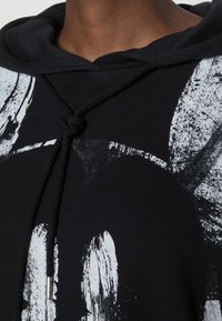 Sudadera negra con cordón, que presenta un diseño gráfico en blanco. El material es suave y el patrón muestra elementos de pinceladas.