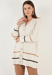 Light beige cardigan with vertical stripes in brown and black. Features a V-neck, long sleeves, and six round buttons down the front.